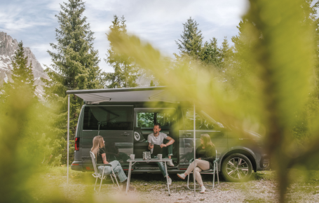 rigofree: Team Coaching und gezielte Schulung von einzelnen Mitarbeitenden beim Business outdoor Coaching in den Dolomiten Das Bild zeigt Paul Felix Rigo beim Coaching mit zwei Angestellten einer Firma. Sie sitzen vor dem VW-Bus an einem Campingtisch. Im Hintergrund sieht man schöne grüne Nadelbäume und die Dolomiten.